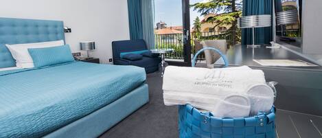 A hotel room with a large bed, a chair, a small table, and a blue basket with towels.