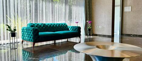 Un hall d’entrée moderne avec un canapé de couleur turquoise, une table basse aux reflets métalliques et une grande fenêtre ornée de rideaux légers.
