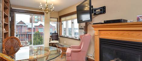 Una sala de estar con una mesa de vidrio, sillas de mimbre, un sillón rosa, una chimenea y un televisor montado.