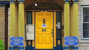 Ein Gebäude mit einer gelben Tür, die mit der Nummer 20 beschriftet ist, flankiert von zwei Säulen und einem Giebelfeld. An beiden Seiten der Tür befinden sich Schilder und vor dem Eingang eine Fußmatte.