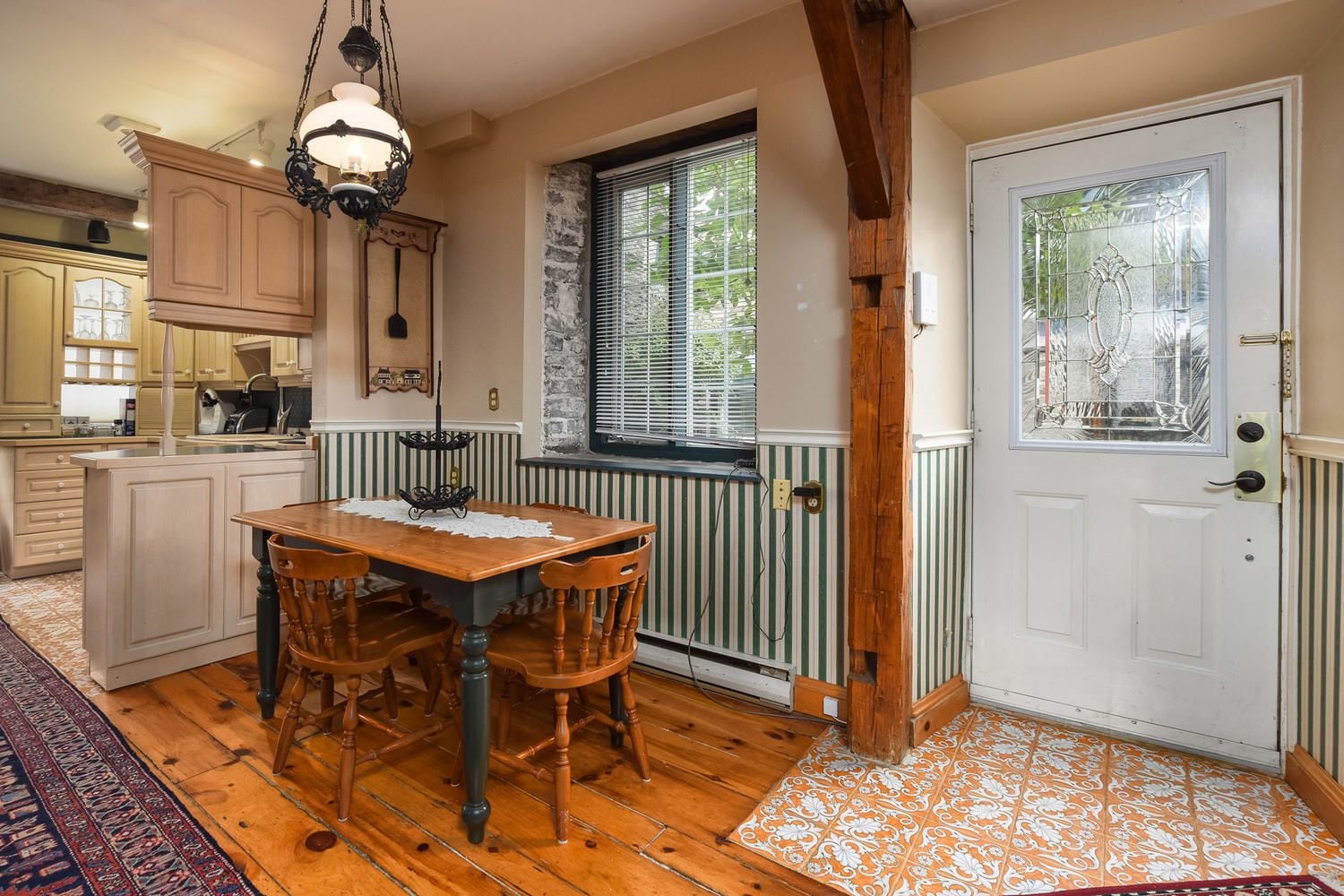 Una cocina con armarios de madera, una mesa de comedor con sillas, una lámpara colgante, un ventanal con persianas y una puerta con panel de vidrio.