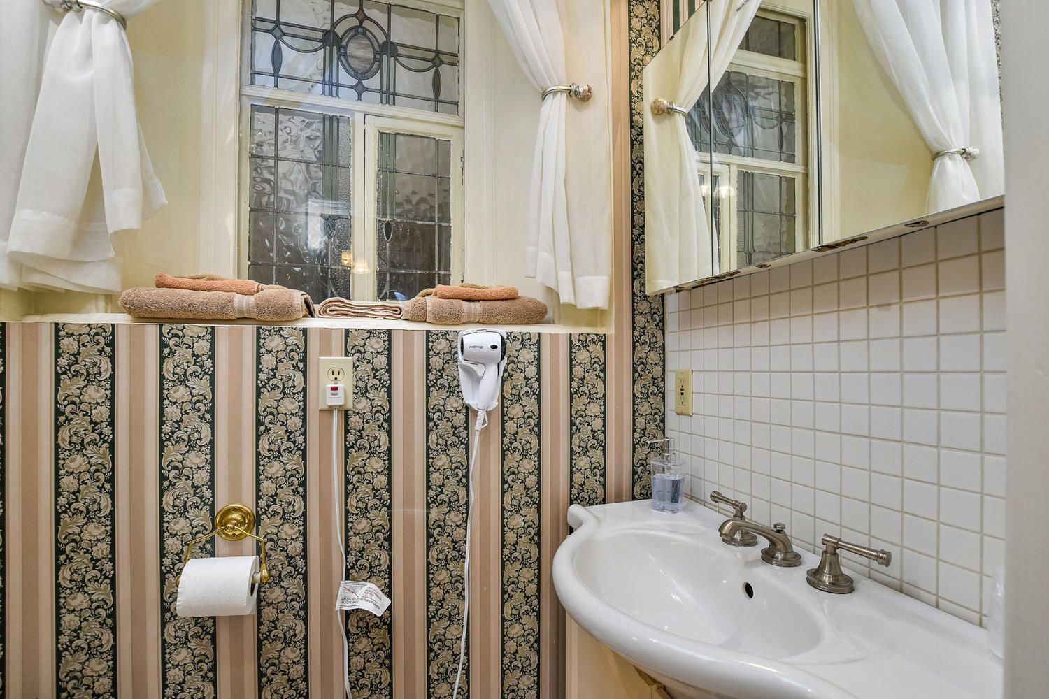 Un baño con lavamanos, porta papel higiénico, perchero para toallas y ventana con cortinas.