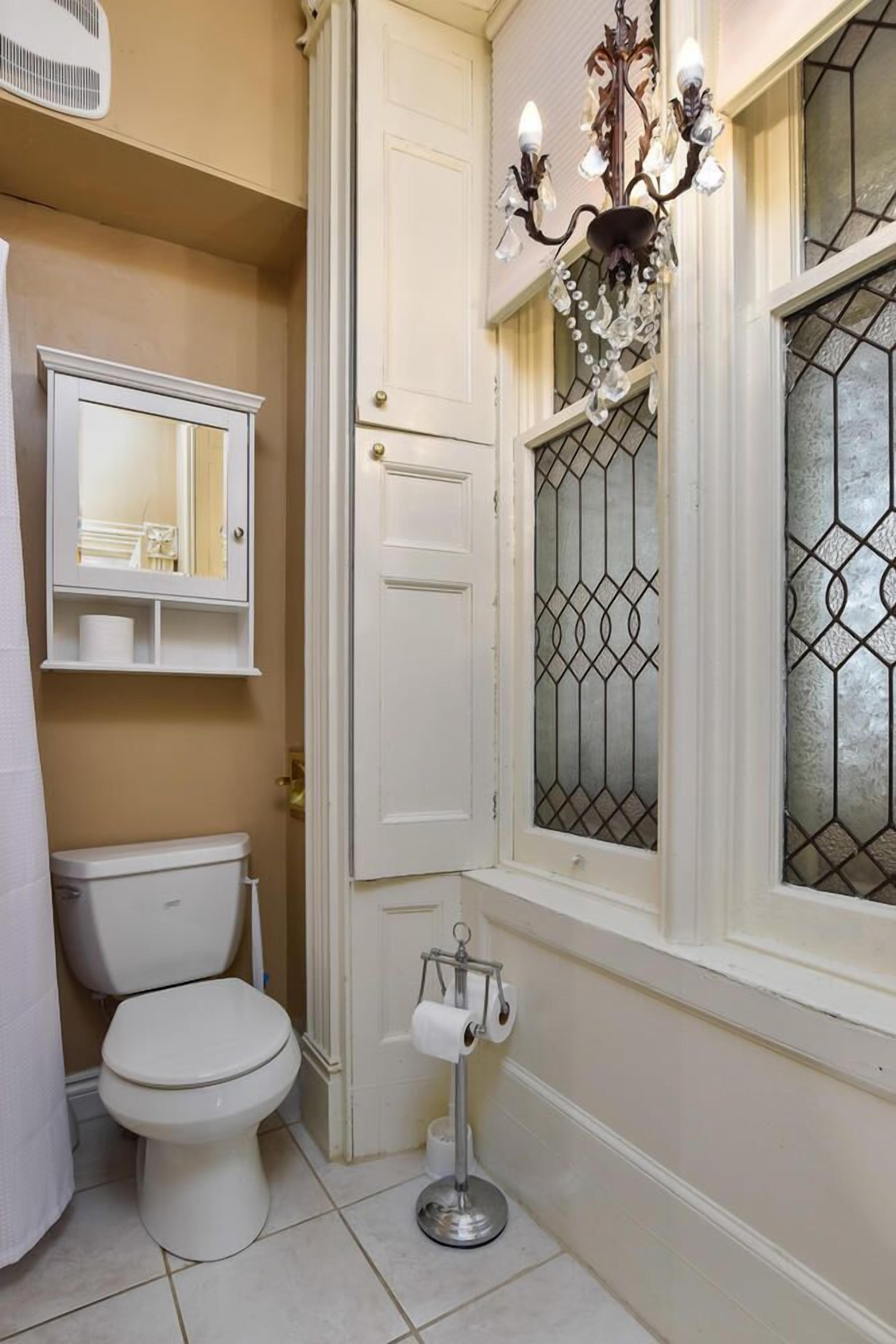 A bathroom with a toilet, a white cabinet with a mirror, a chandelier, and a window with decorative glass.