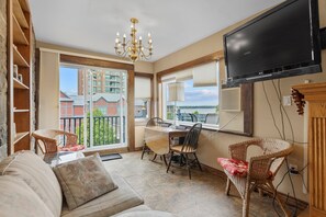 A living room with a sofa, dining table, chairs, a chandelier, a TV, and a view of buildings and water outside.