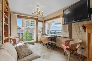 Una sala de estar con sofá, mesa de comedor, sillas, candelabro, televisor y vistas de edificios y agua al exterior.