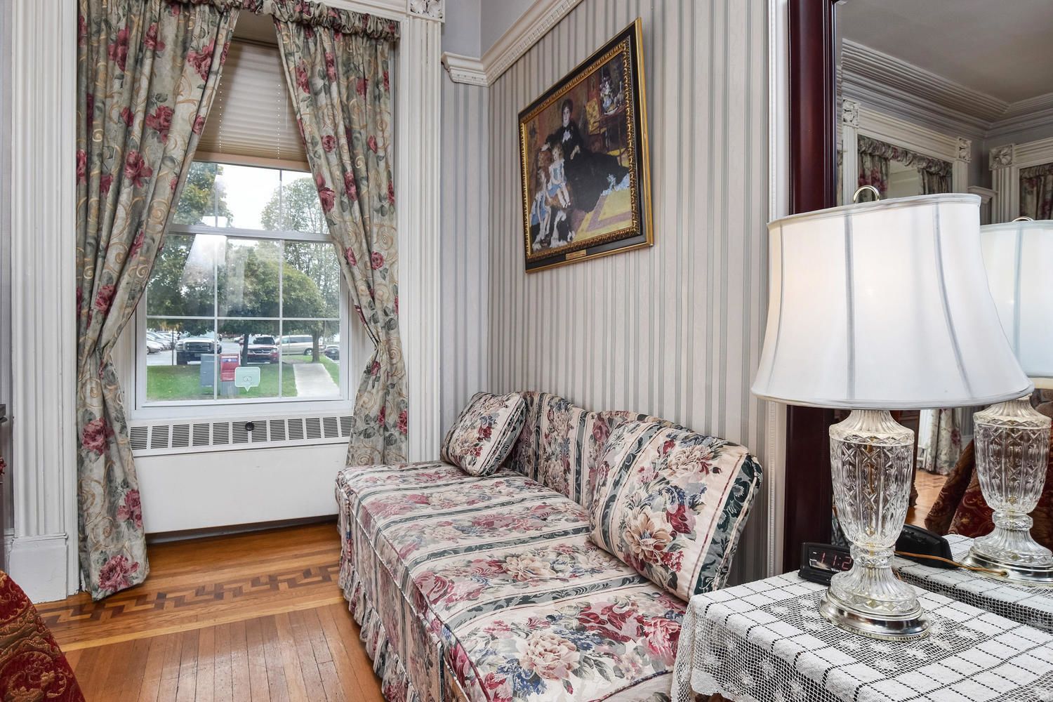 A room with a floral-patterned sofa, a table with a lace tablecloth, and a lamp with a white shade.