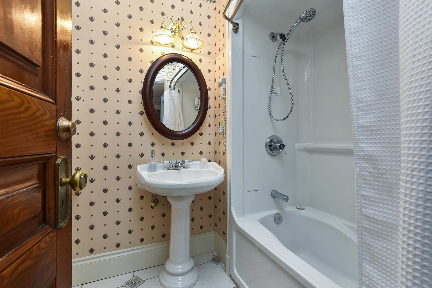 A bathroom with a wooden door, a white sink with a mirror, patterned wallpaper, and a white bathtub with a shower head.