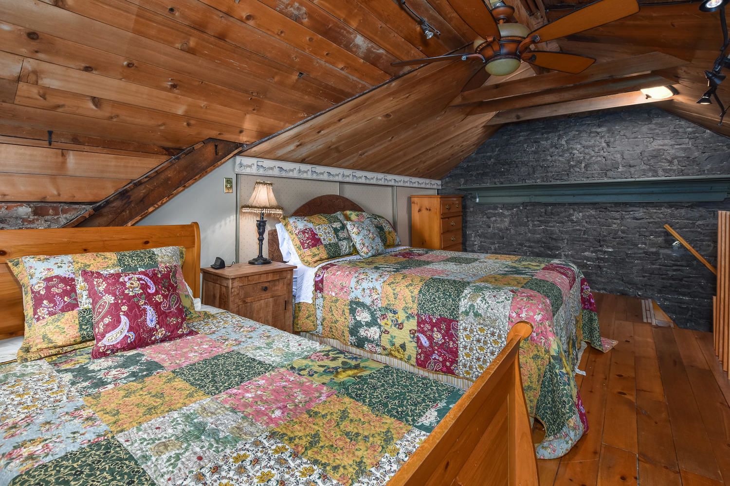Une chambre confortable située sous les toits, avec deux lits en bois, un ventilateur de plafond en bois et un mur en pierre.