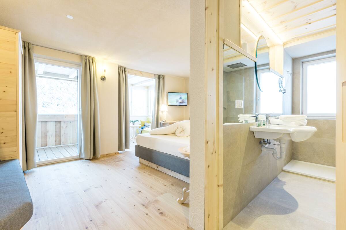 A modern hotel room with a wooden floor, a bed, a bathroom with a sink and mirror, and a balcony with curtains.