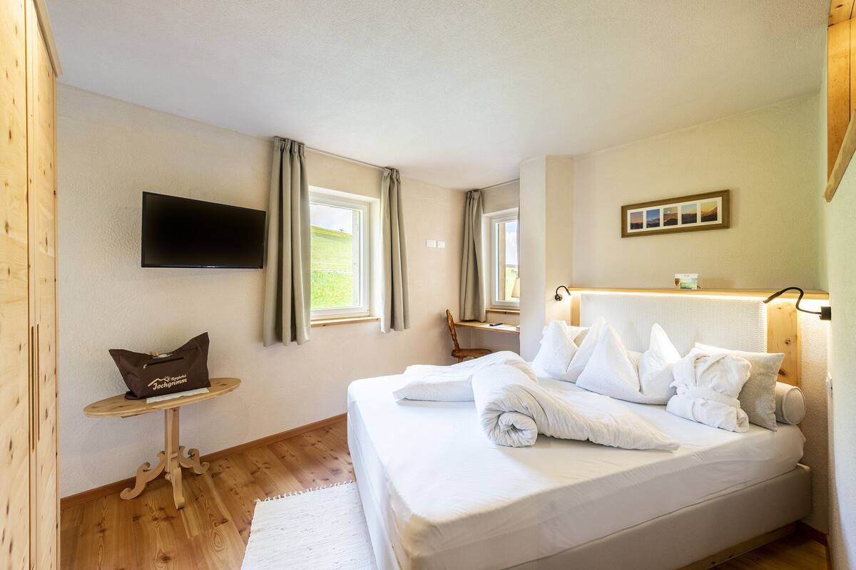 A neatly made bed with white linens, a small wooden table with a bag, and a TV mounted on the wall.
