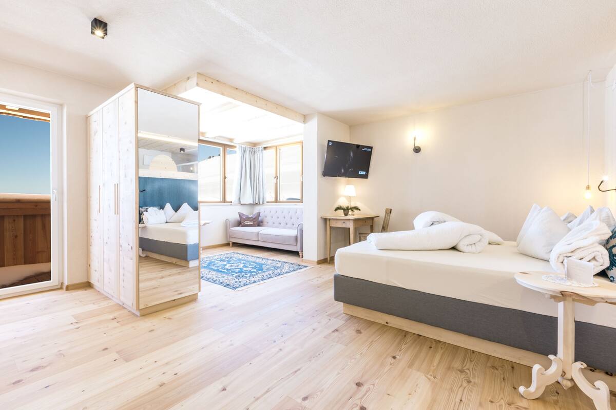 A modern hotel room with a wooden floor, a bed with white linens, a sofa, a TV mounted on the wall, and a small table with a lamp.