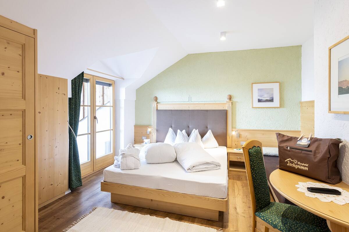 A hotel room with a wooden bed, a green chair, a small table, and a wooden wardrobe.