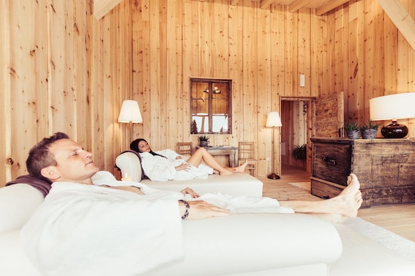 A wooden cabin interior with two people relaxing on a couch, a wooden dresser, and a window with a view. - Berghotel Jochgrimm - Your hoome in the Dolomites (Aldino)