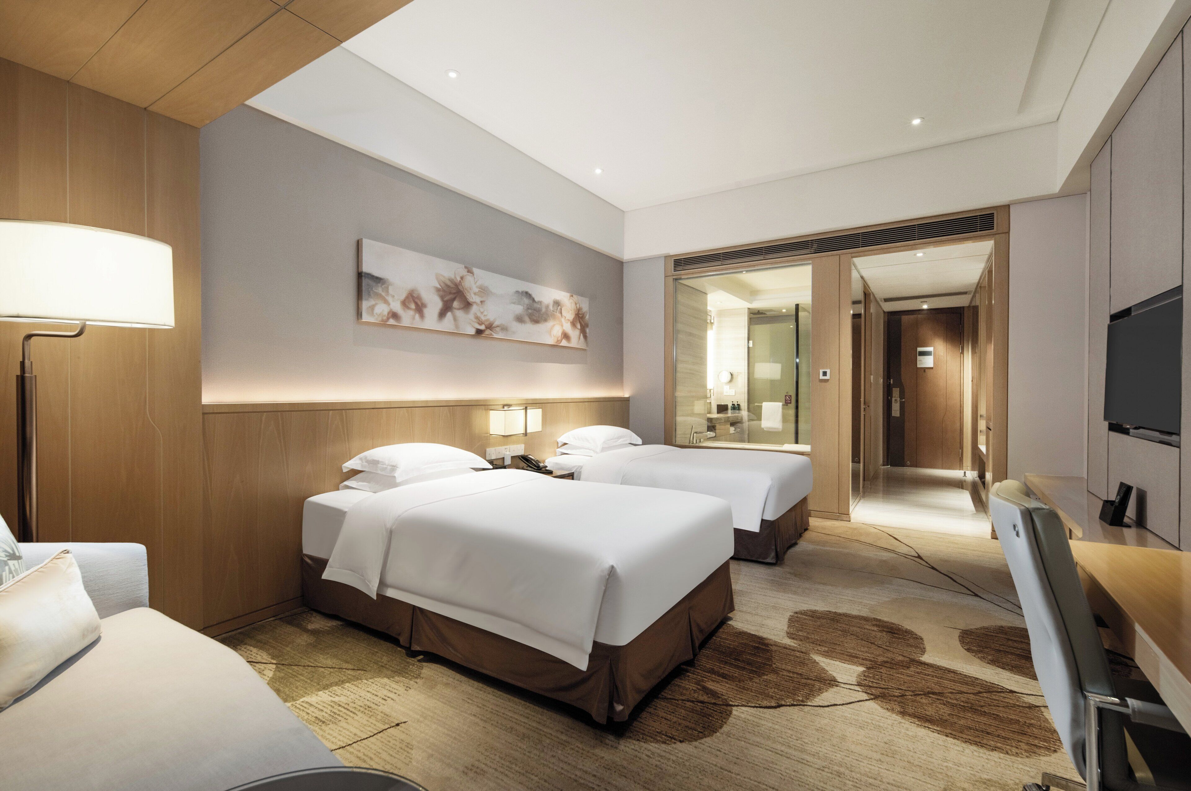 A hotel room with two beds, a desk, a lamp, and a painting on the wall.