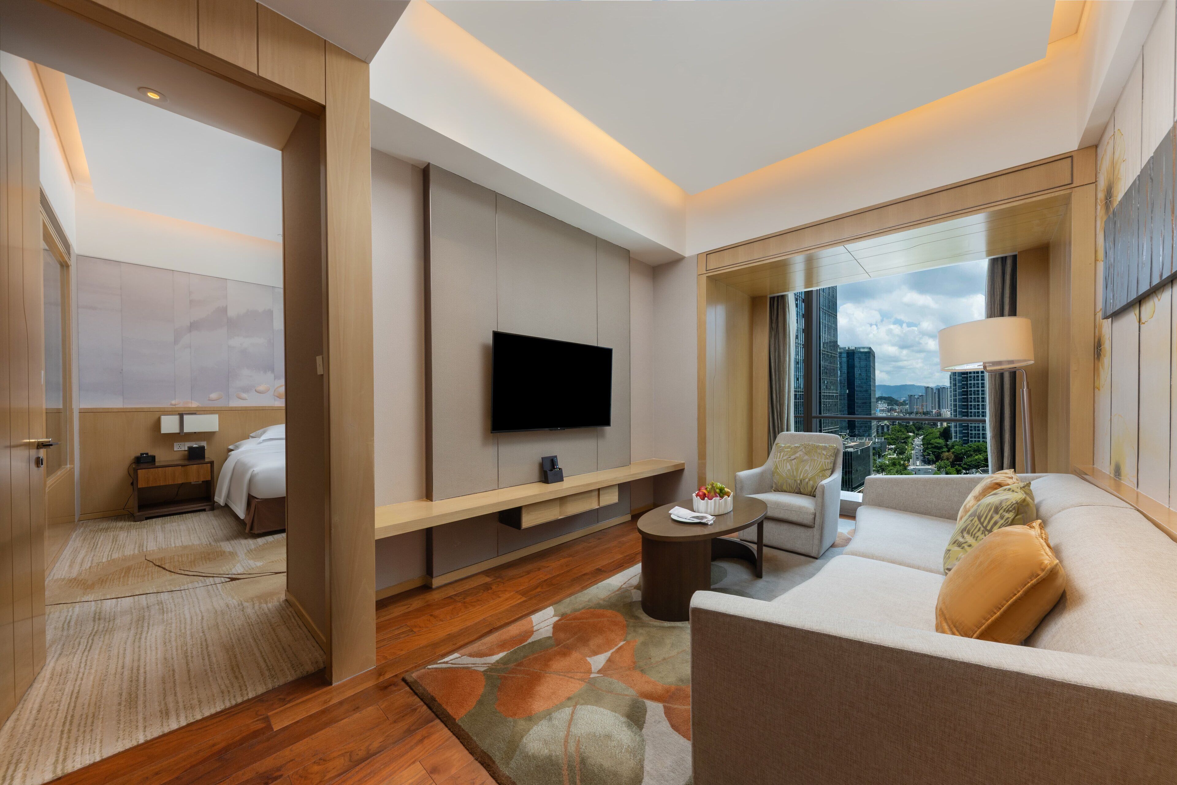 A modern hotel room with a large TV, a sofa, a coffee table, and a city view.