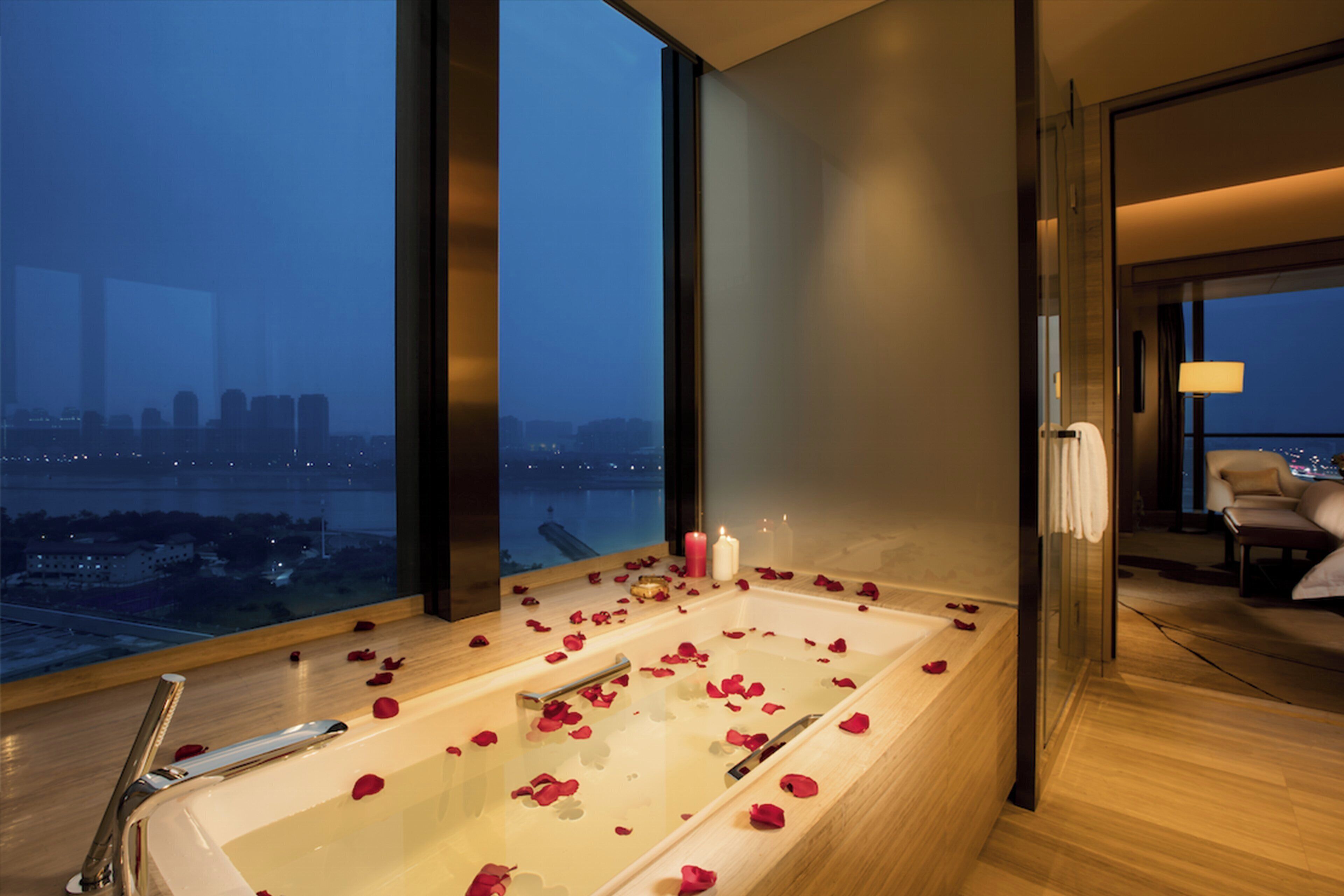 A modern bathroom with a large bathtub filled with rose petals, a city view, and a city skyline visible through the window.