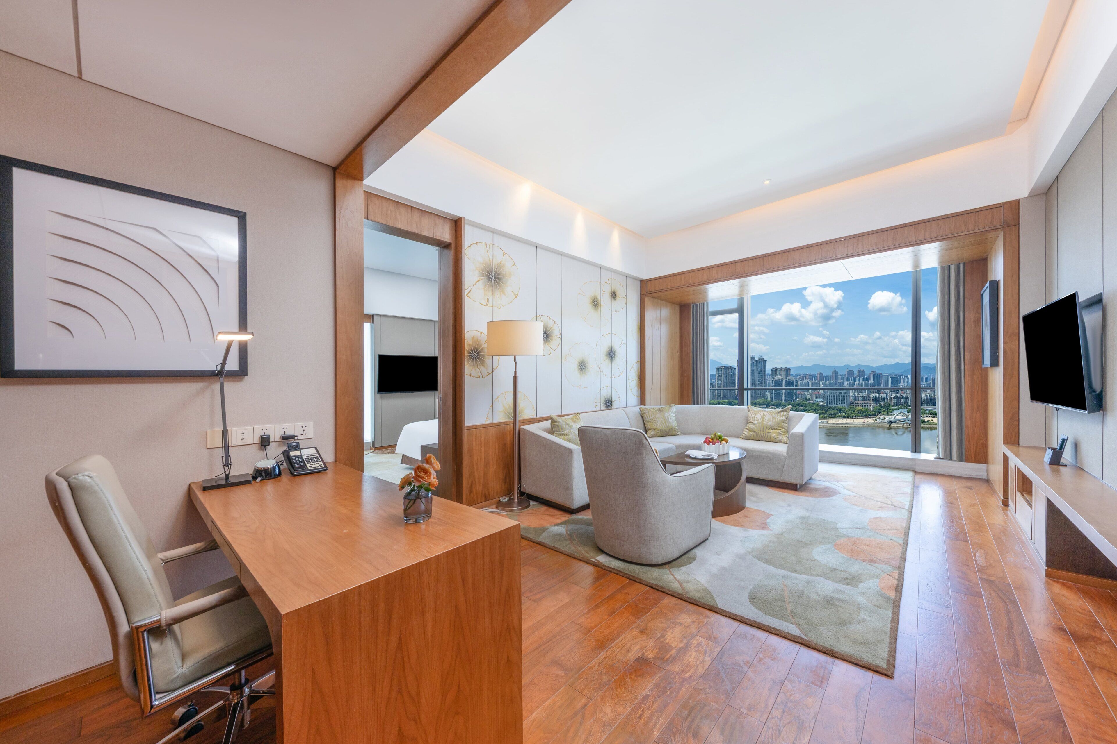Ein modernes Hotelzimmer mit Schreibtisch aus Holz, einer Couch, einem großen Fenster mit Blick auf die Stadt und einem Flachbildfernseher.