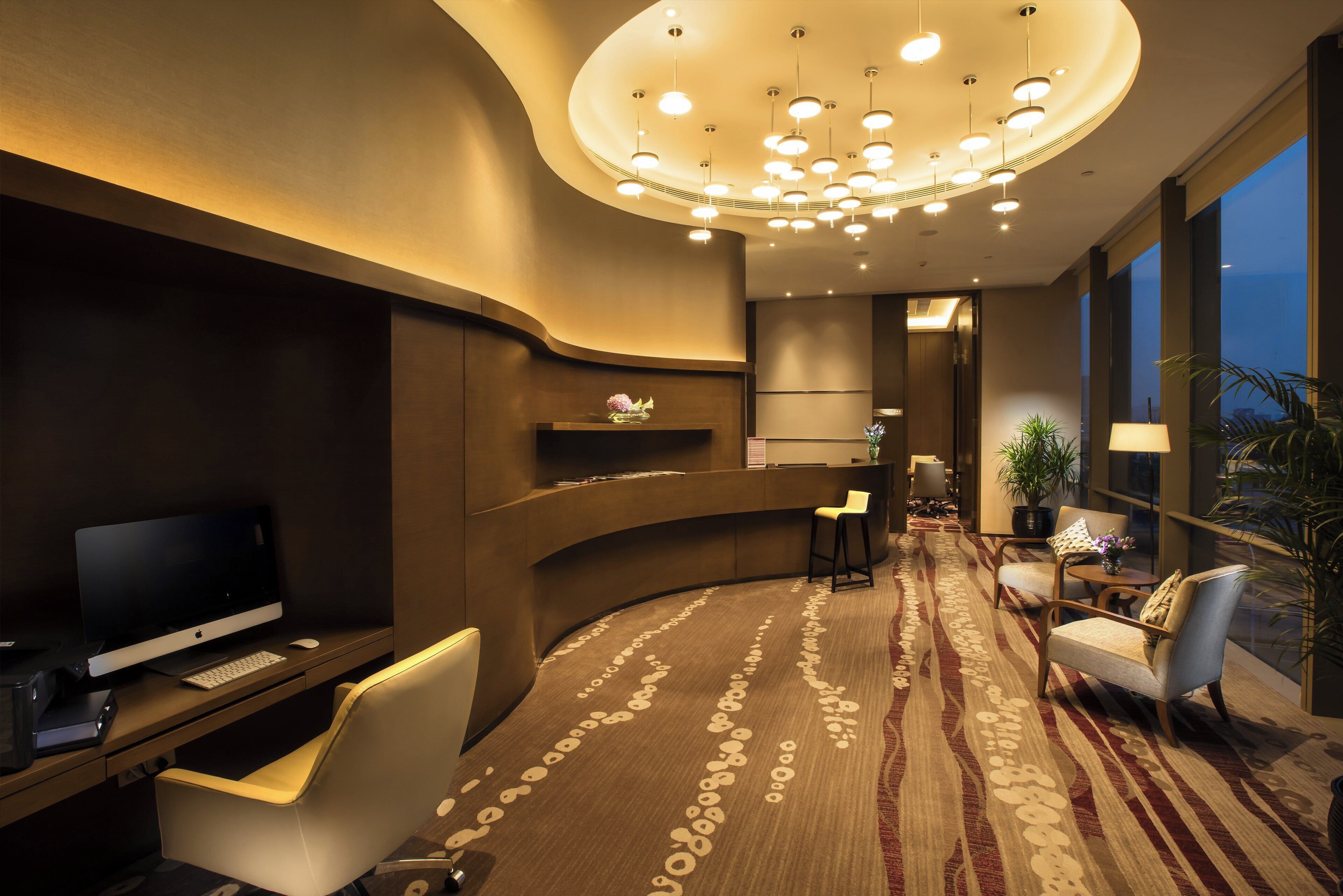 A modern hotel lobby with a curved reception desk, a flat-screen TV, seating area, and a large window offering a view.