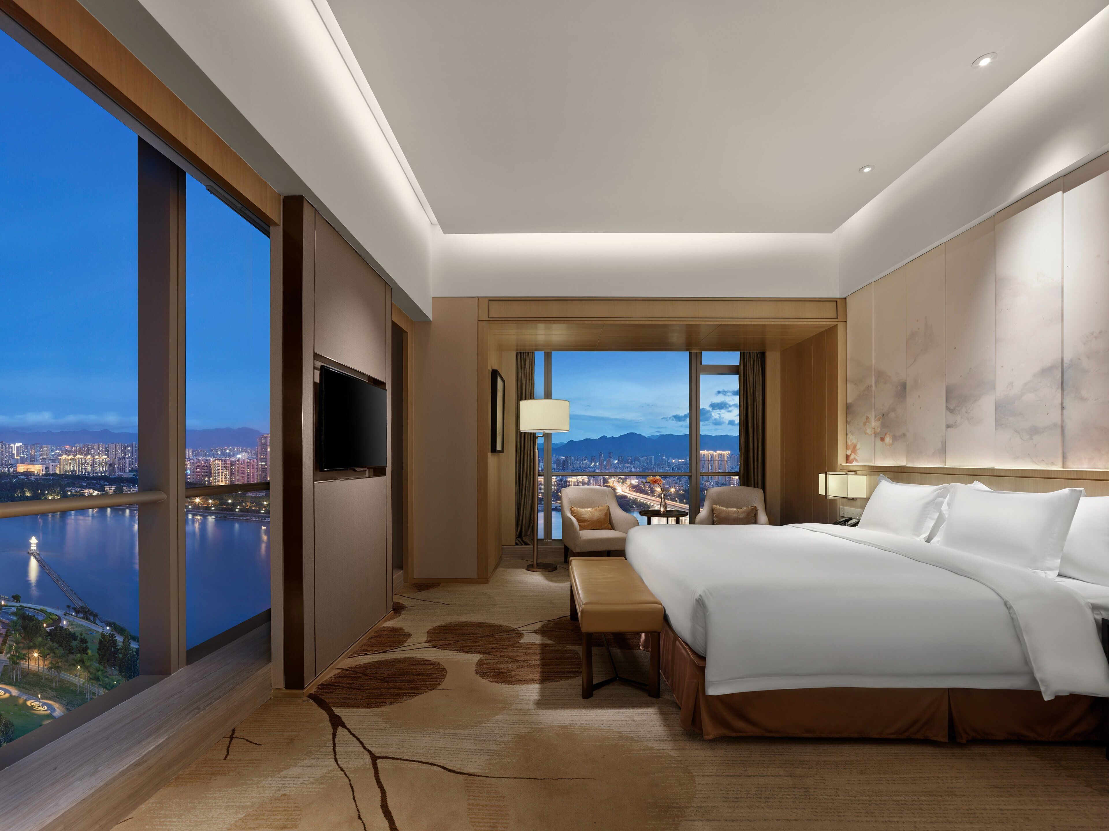 A hotel room with a large bed, a desk, a chair, and a TV. There is a view of a cityscape and a body of water through large windows.