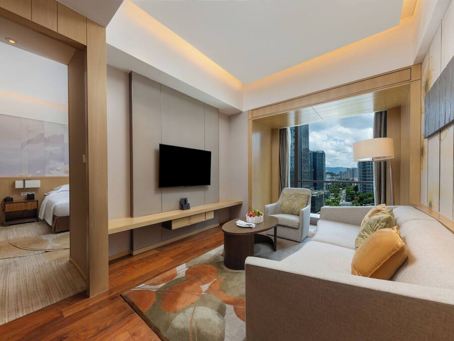 A modern hotel room with a flat-screen TV, a sofa, a coffee table, and a city view through large windows.