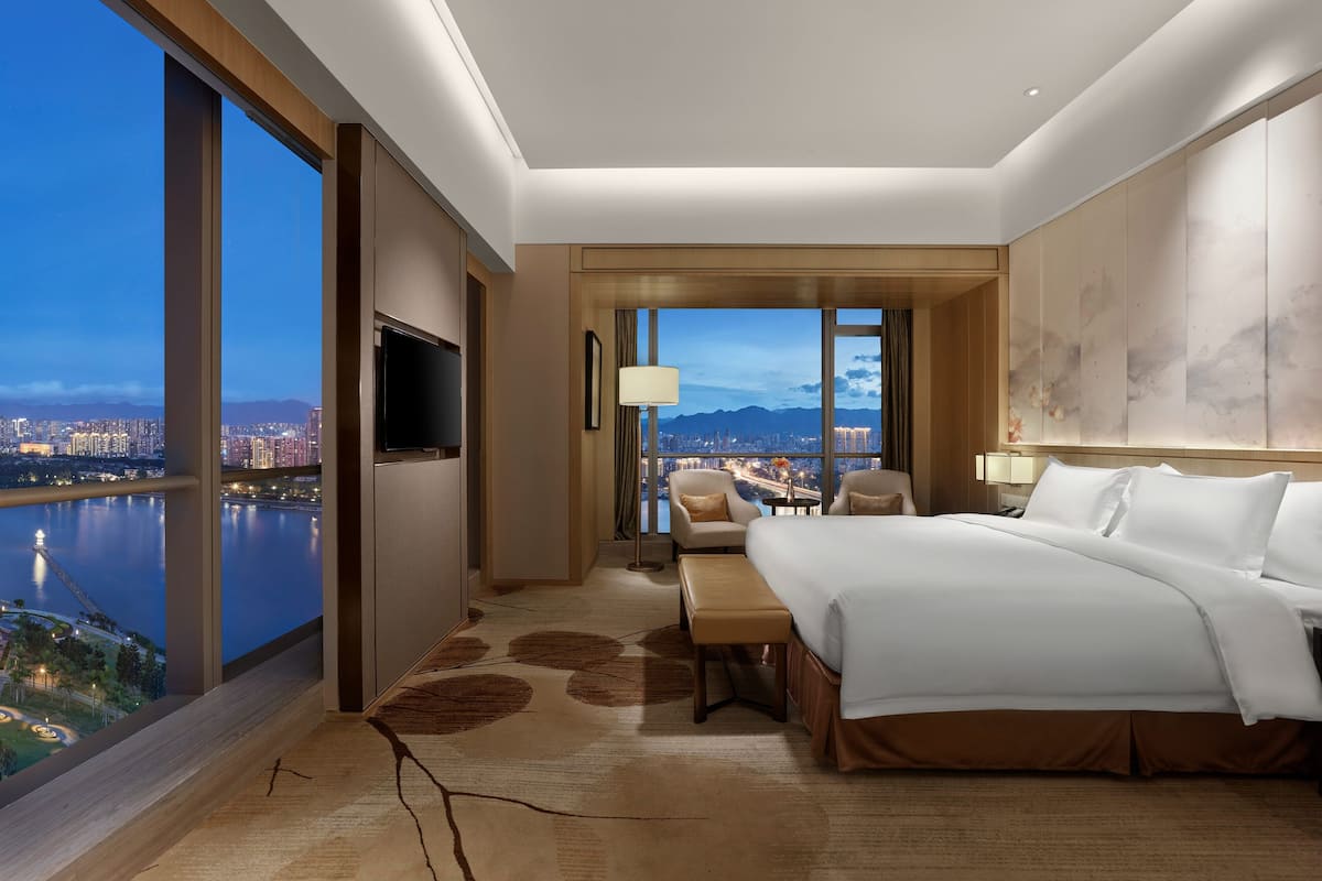 Ett modernt hotellrum med en stor säng, en TV, ett skrivbord med en stol och utsikt över en stad och berg.