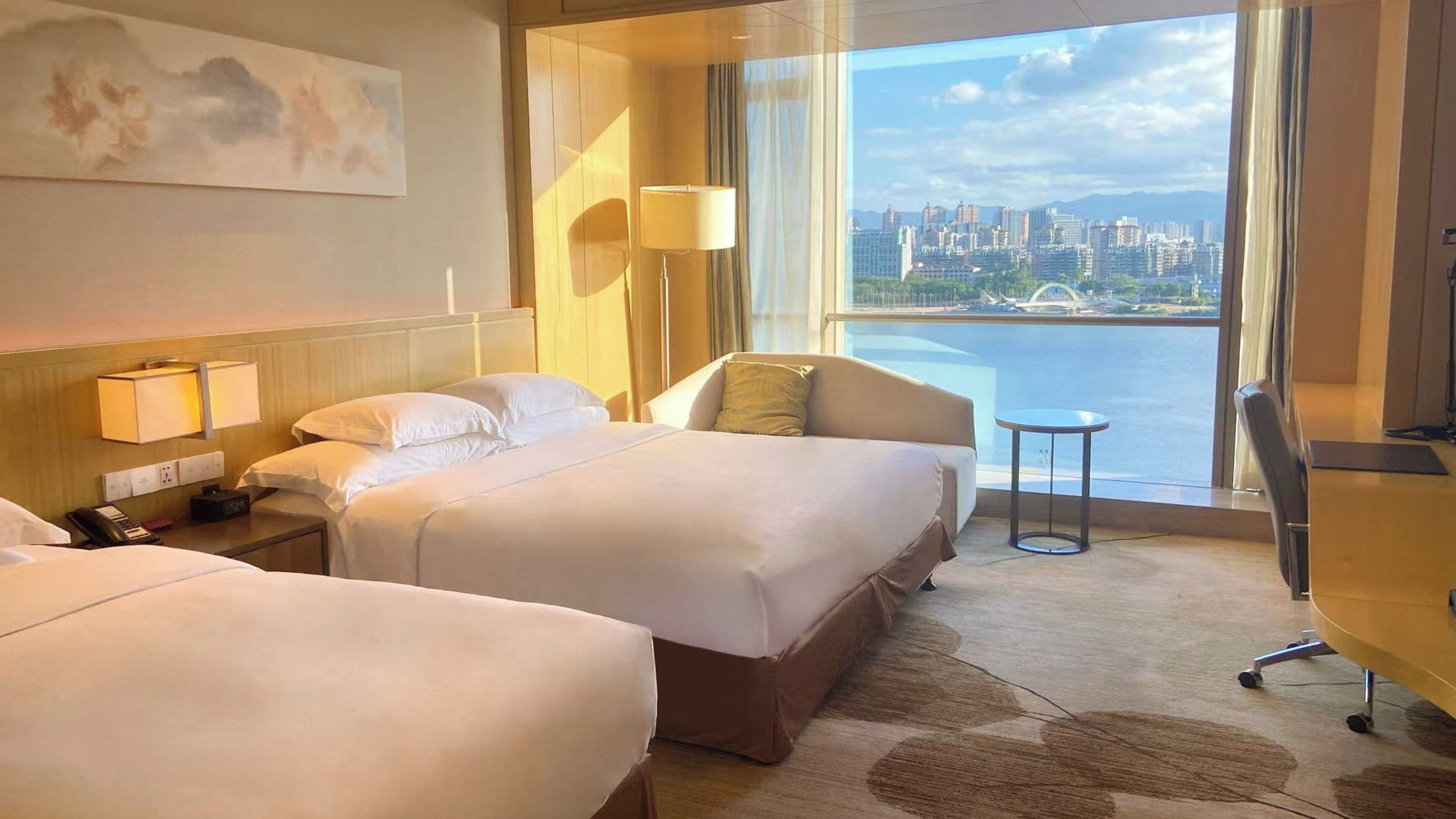 Habitación de hotel con dos camas, un escritorio, una silla y vistas a la ciudad y al agua.