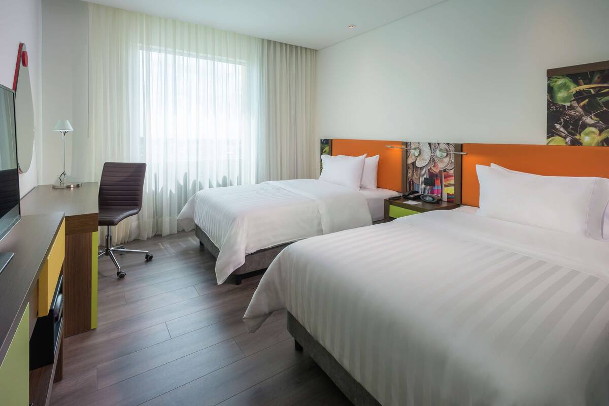 Habitación de hotel con dos camas, un escritorio, una silla y un gran ventanal con cortinas.