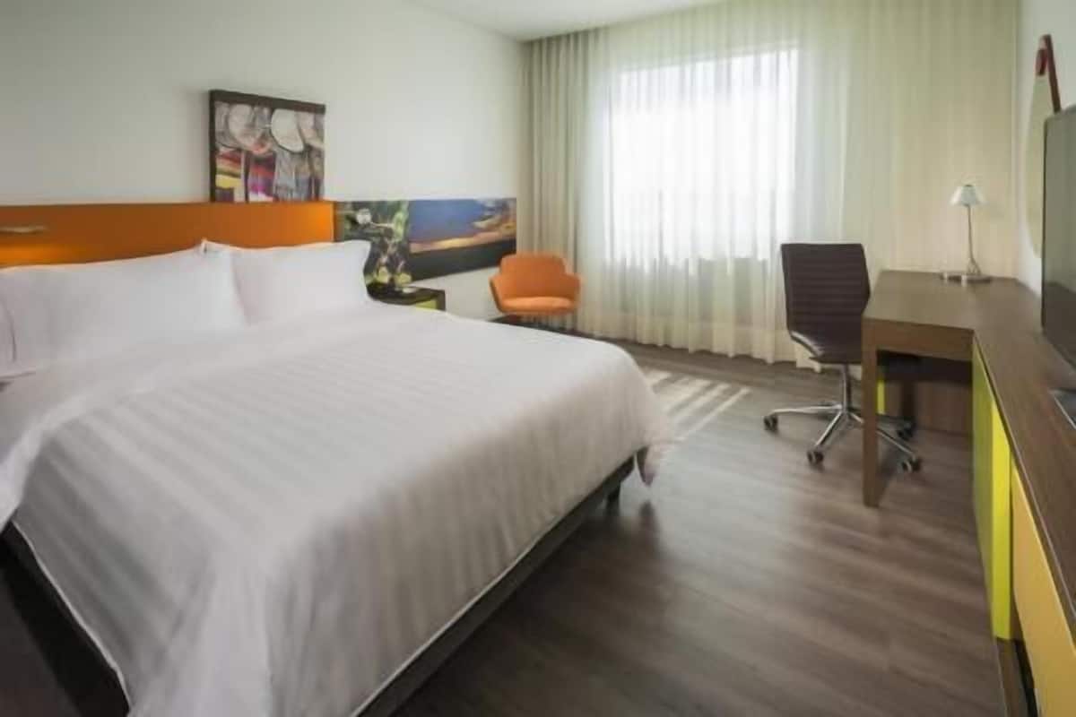 Habitación de hotel con una cama grande, un escritorio, una silla de oficina, un cuadro en la pared y una ventana con cortinas.