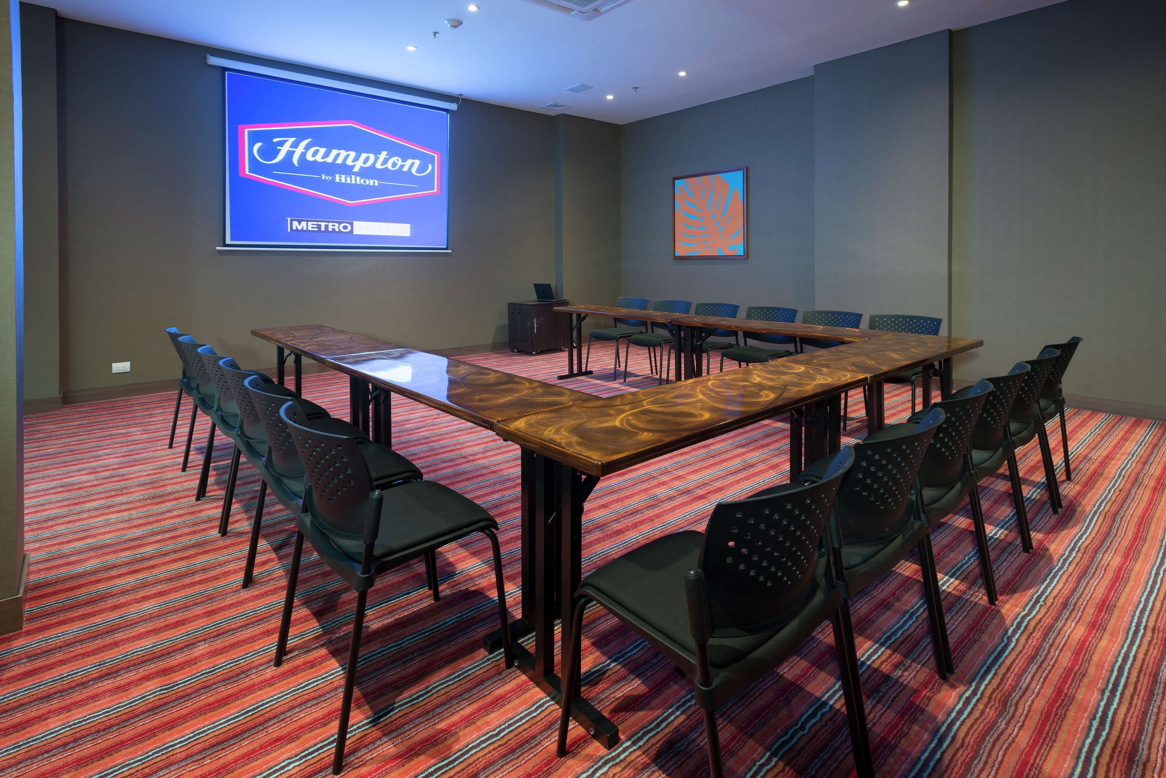 A conference room with a long table, chairs, a projector screen displaying 'Hampton by Hilton - METRO', and a patterned carpet.