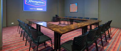 A conference room with a long table, chairs, a projector screen displaying 'Hampton by Hilton - METRO', and a patterned carpet.