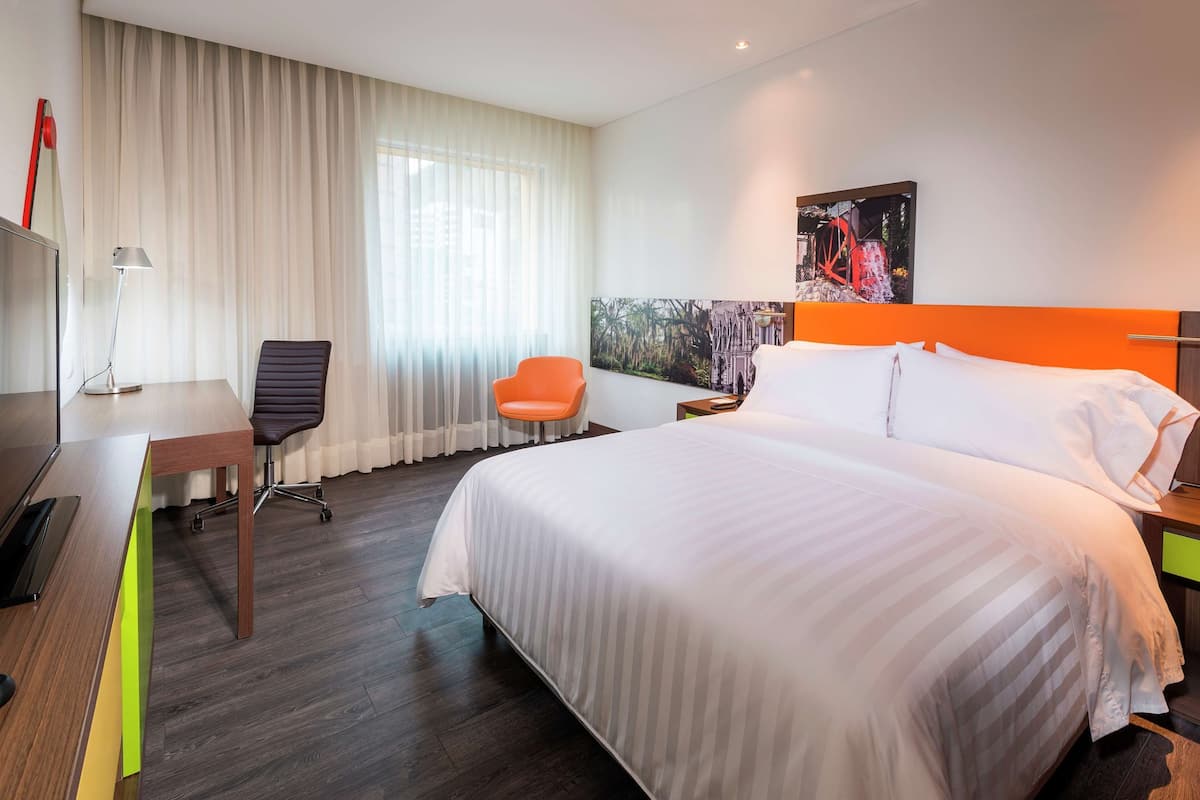 Habitación de hotel con cama, escritorio, silla y un amplio ventanal con cortinas.
