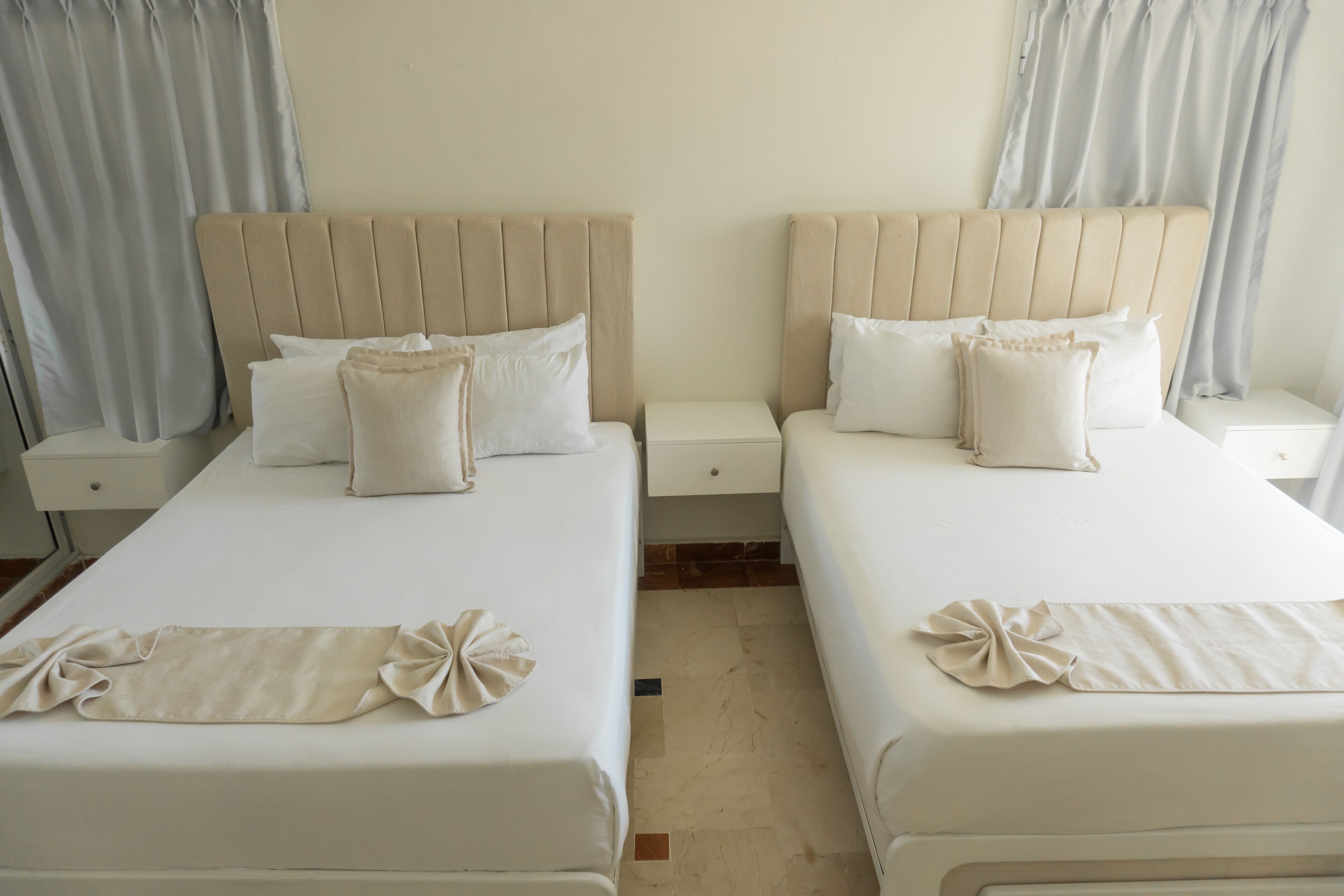A hotel room with two beds, each with a headboard, white bedding, and a small bedside table.