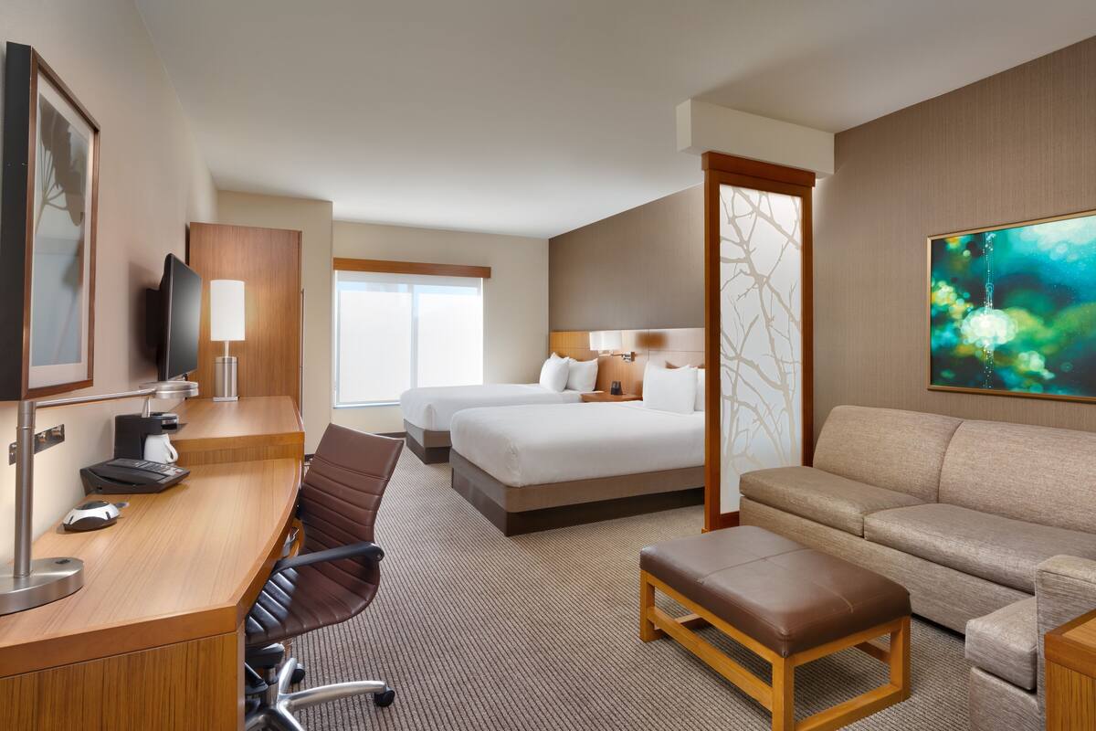 A hotel room with a desk, two beds, a sofa, and a large window.