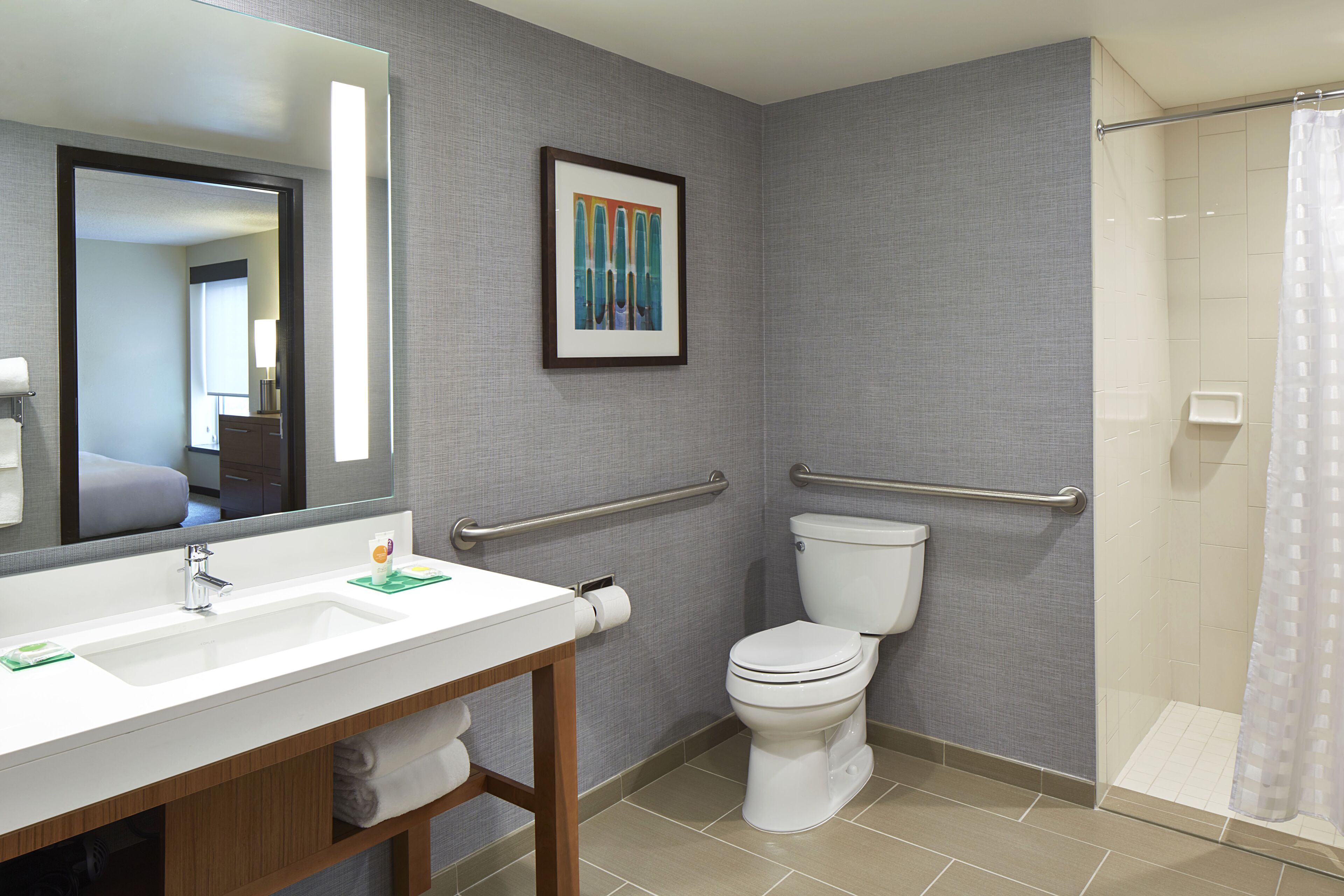 Un baño moderno con lavamanos, espejo y retrete blancos, que incluye una ducha con puerta de cristal y barra de apoyo.