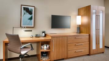 A hotel room with a wooden desk, a chair, a flat-screen TV, a lamp, a minibar, and a wardrobe.
