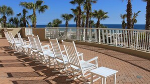 A row of white rocking chairs and a table on a brick patio with a view of the ocean and palm trees. - Indigo by Luxury Coastal Vacations (Pensacola)