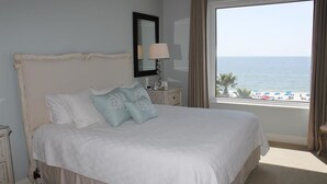 A bedroom with a large bed, a nightstand with a lamp, a mirror, and a view of the beach. - Indigo by Luxury Coastal Vacations (Pensacola)