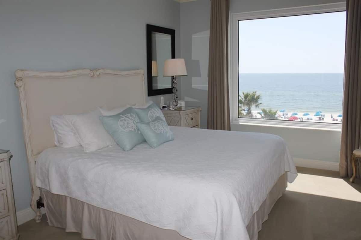A bedroom with a large bed, a nightstand with a lamp, a mirror, and a view of the beach.