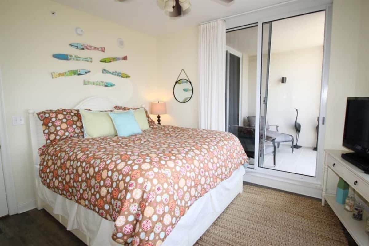 A bedroom with a bed, a TV, a lamp, and a sliding glass door leading to a balcony.