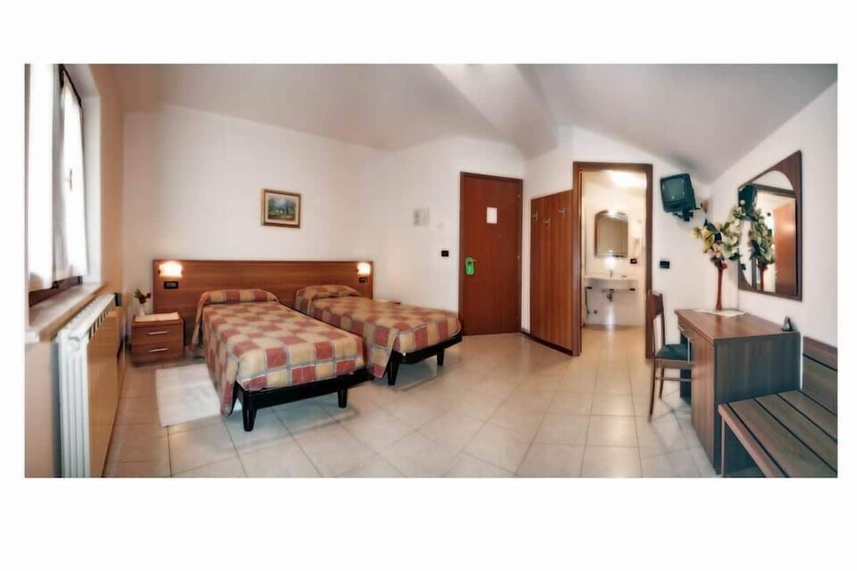 A hotel room with two beds, a wooden headboard, a small bedside table, a nightstand, a bathroom with a mirror, a sink, and a wooden bench.