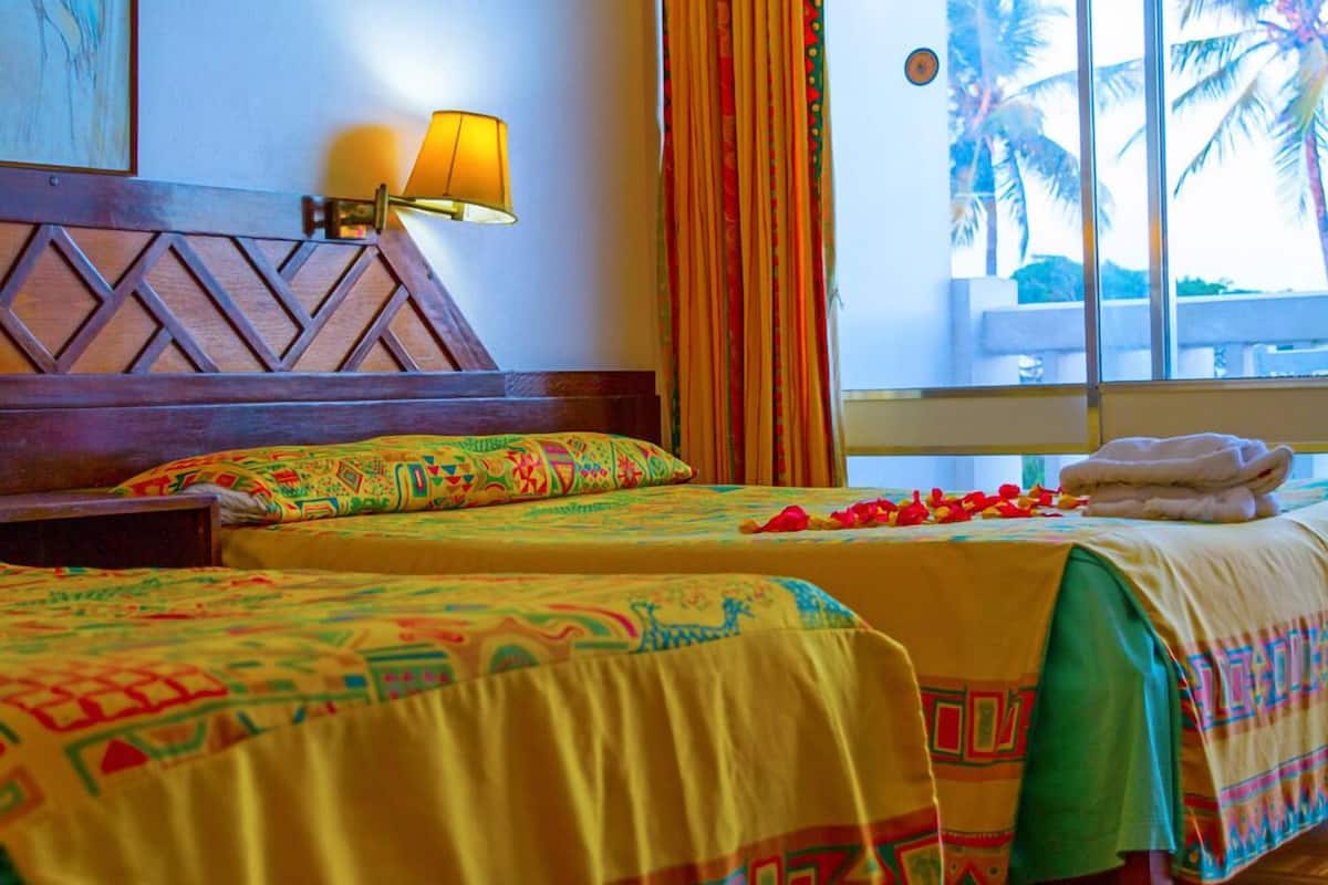A hotel room with two beds, a wooden headboard, a nightstand, a lamp, a window with curtains, and a view of palm trees.