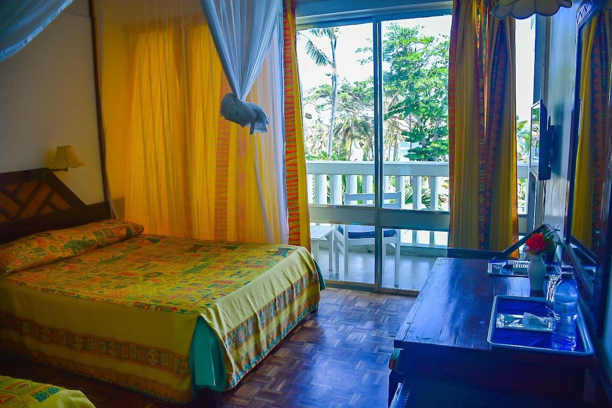 A room with a bed, a desk with a mirror, and a balcony with a view of palm trees.