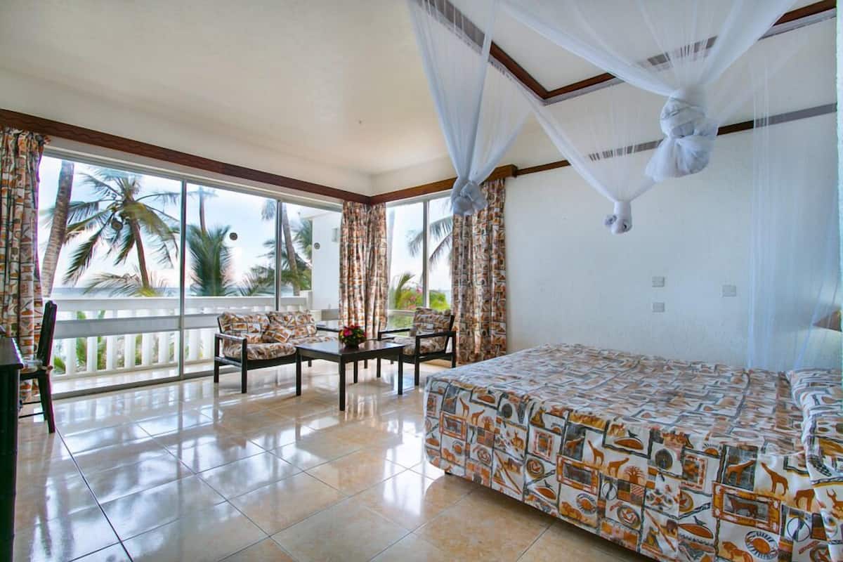 A hotel room with a bed, a balcony with a view of palm trees, and a dining area with a table and chairs.