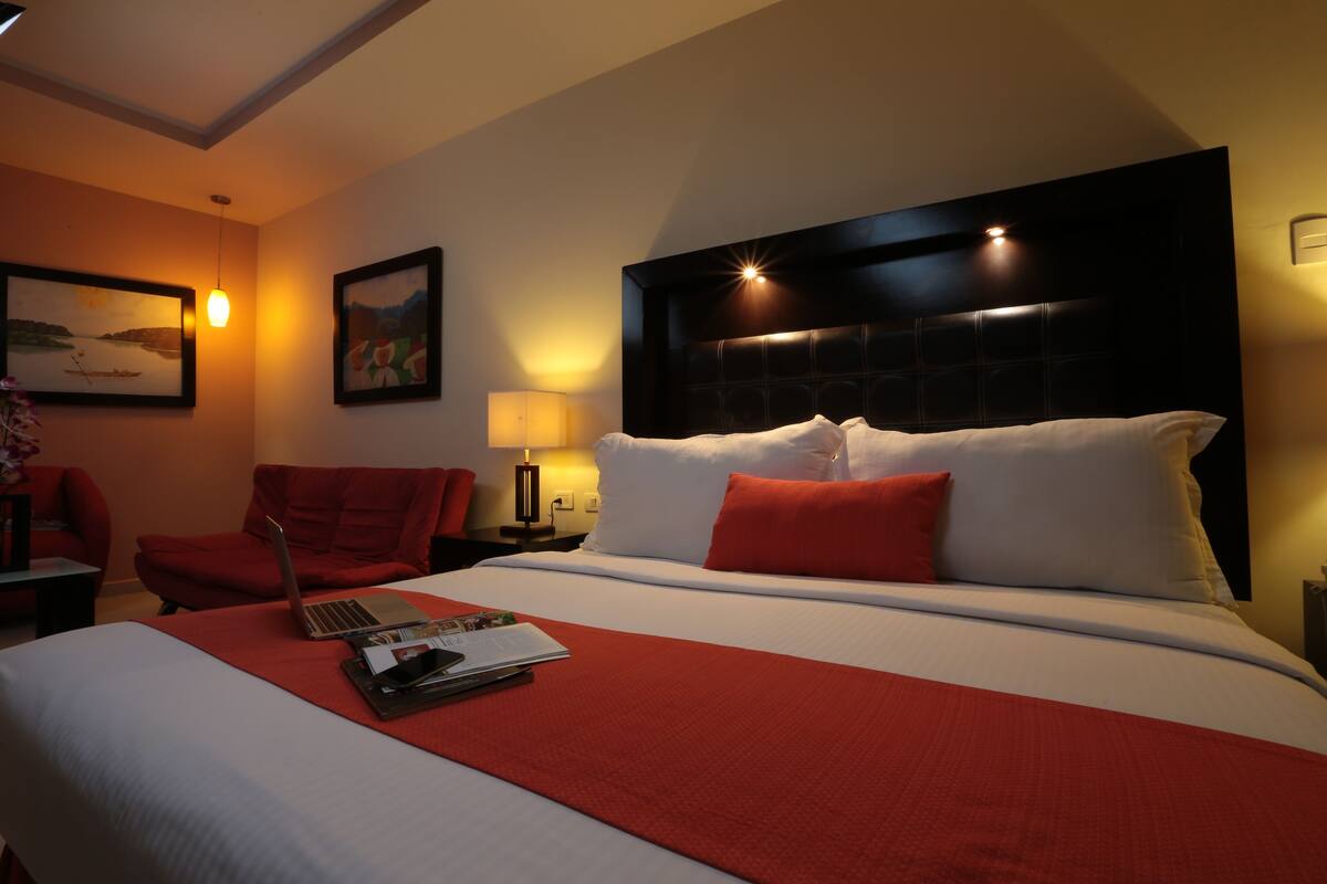 A hotel room with a large bed, a desk with a laptop, a chair, and a nightstand with a lamp.