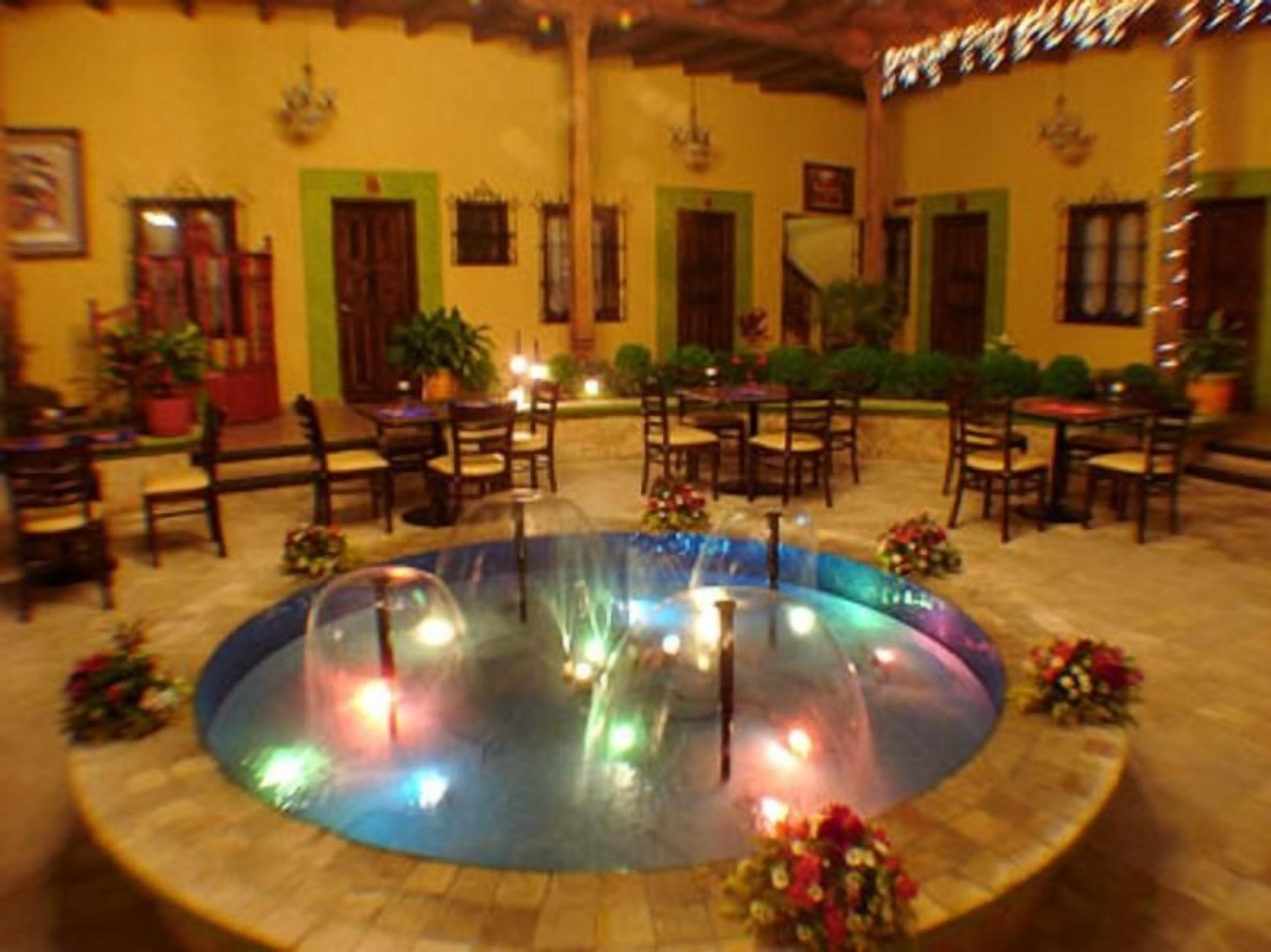 Une fontaine circulaire illuminée, entourée de tables et de chaises, se trouve dans une cour agrémentée de plantes en pot et bordée d’un bâtiment aux portes vertes.
