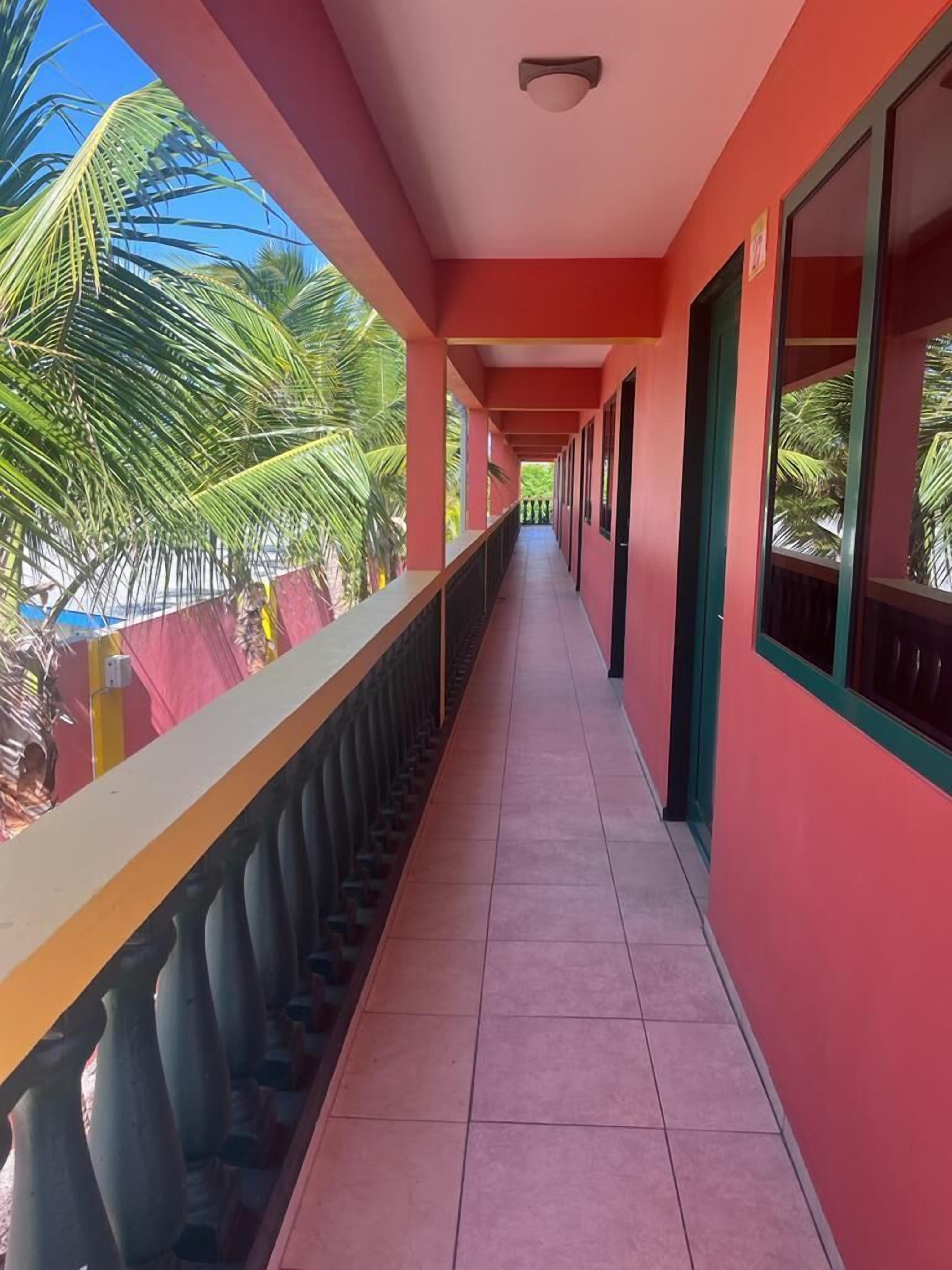 A corridor with red walls and a tiled floor, leading to a balcony with a view of palm trees.