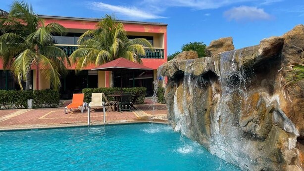 A swimming pool with a waterfall feature, surrounded by palm trees and a building with balconies. - Del Rey Apartments (Noord)