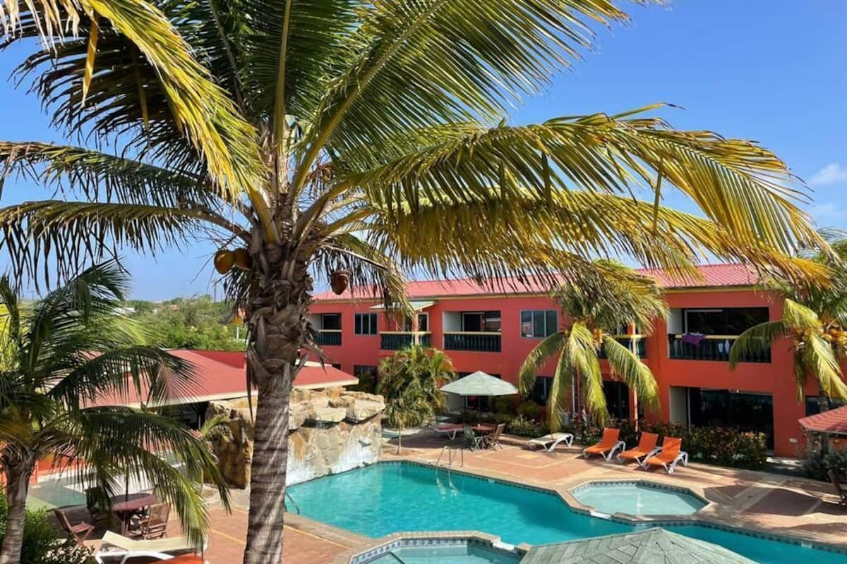 A resort with a swimming pool, palm trees, and a red building with balconies.
