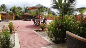 An outdoor patio area with brick flooring, wooden tables, and chairs, surrounded by palm trees and flowering plants. - Del Rey Apartments (Noord)