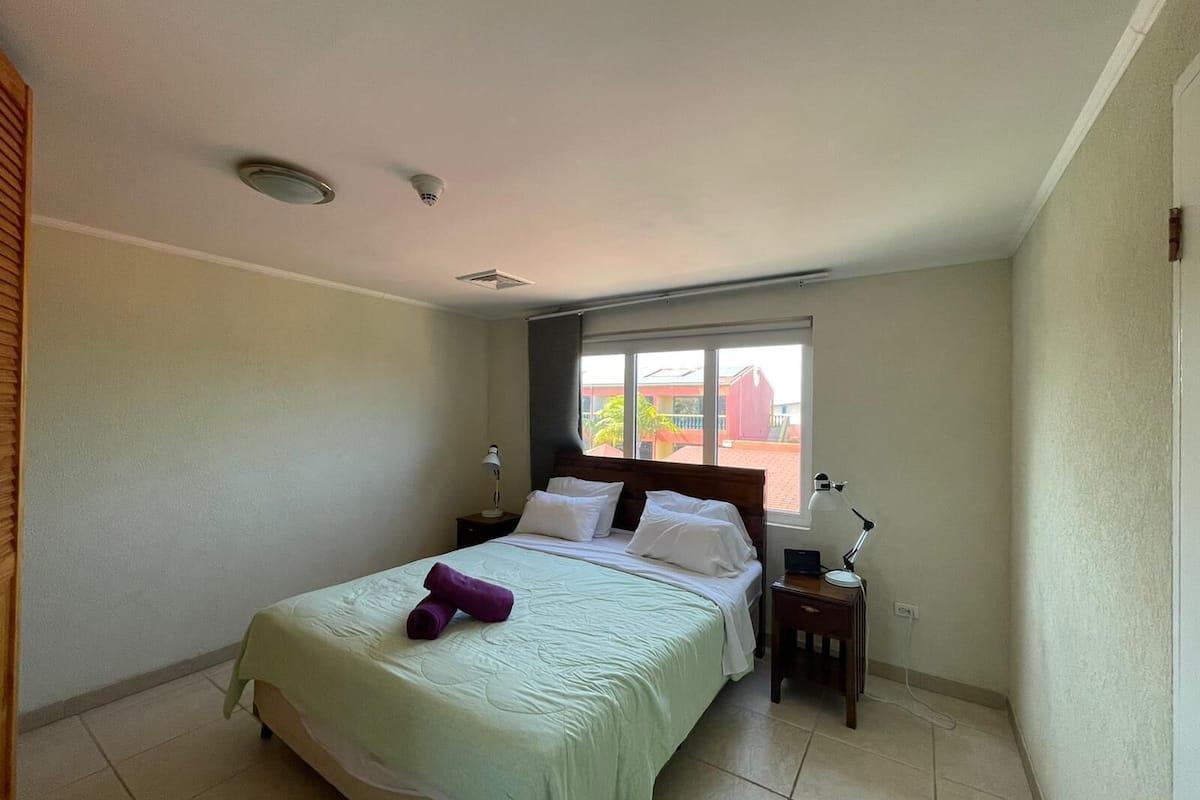 A single bed with a green bedspread, two pillows, and a purple towel. There are two bedside tables with lamps. A window with a view of buildings is visible.